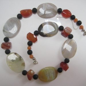 Zu einer Schnecke gelegte Edelsteinkette aus Onyx, Achat und Karneol. Sie besteht aus Achat-Elipsen in verschiedenen Farbtönen mit Onyxkugeln und orangen Karneolnuggets.