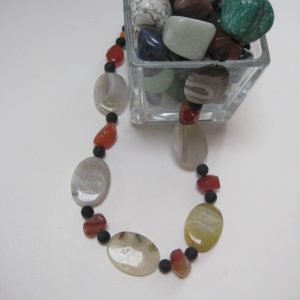 Eine Edelsteinkette aus Onyx, Achat und Karneol, die aus einem Glasbehälter mit Edelsteinen fließt. Sie besteht aus Achat-Elipsen in verschiedenen Farbtönen mit Onyxkugeln und orangen Karneolnuggets.