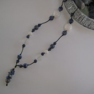 Eine Edelsteinkette aus Sodalith, Koralle und Bergkristall gleitet aus einem Glas mit weiteren Edelsteinen. Zu sehen sind große, polierte Sodalith Kugeln, matte Korallen und weiße Jadelkugeln.