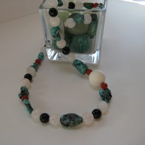 Edelsteinkette aus Türkis, Rosenquarz, Karneol und Onyx auf einer weißen Damenbüste, die aus einem Glasgefäß fließt. Die Steine sind türkis, rose, weiß und schwarz.
